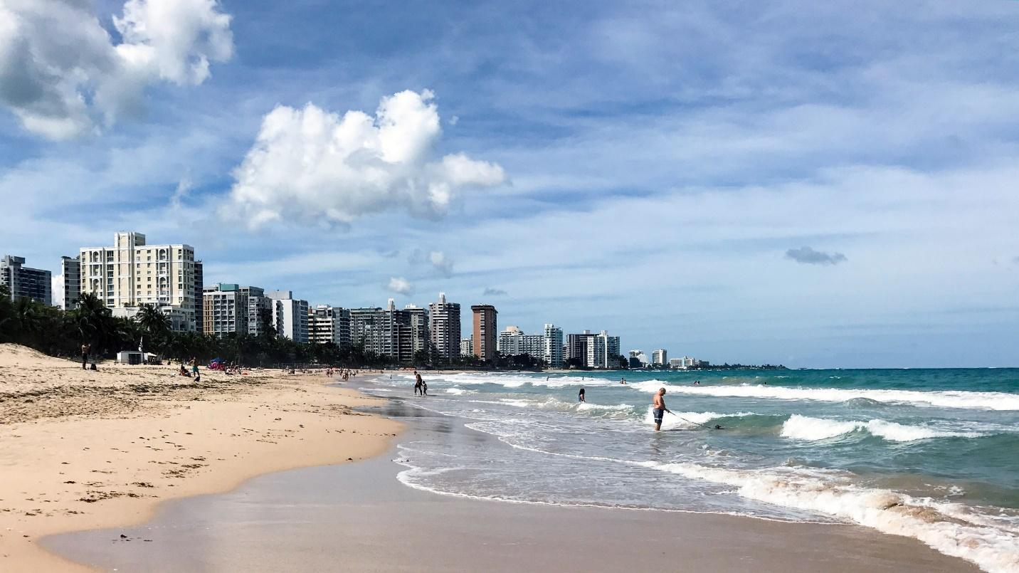 A handful of beachgoers walk through sand and surf with buildings in the background in a section about Ocean Park Beach in the San Juan area. 