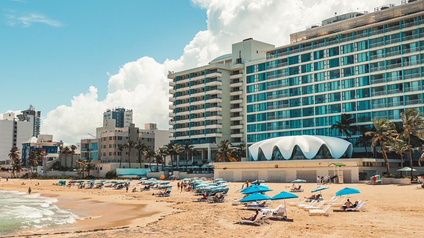 Beachgoers, lounge chairs with umbrellas, and buildings on the sand in a section about Condado Beach. 