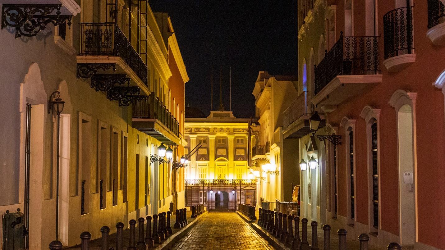 The cobblestone street of Calle de la Fortaleza flanked by buildings at night in a section describing historic Old San Juan, Puerto Rico. 