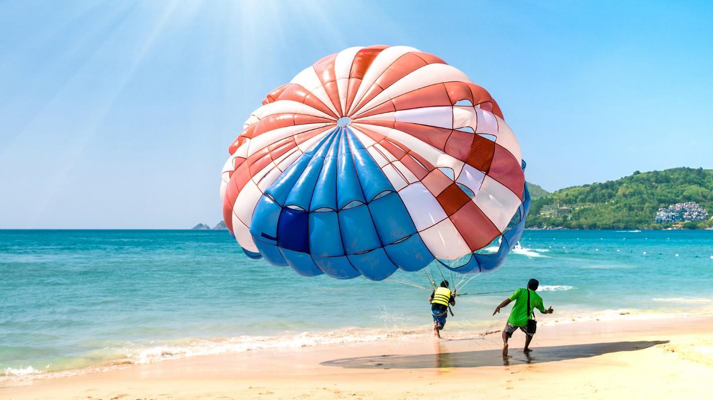 Two people run down the beach with one pulling a red, white, and blue parachute glider in a section about outdoor activities in Condado. 