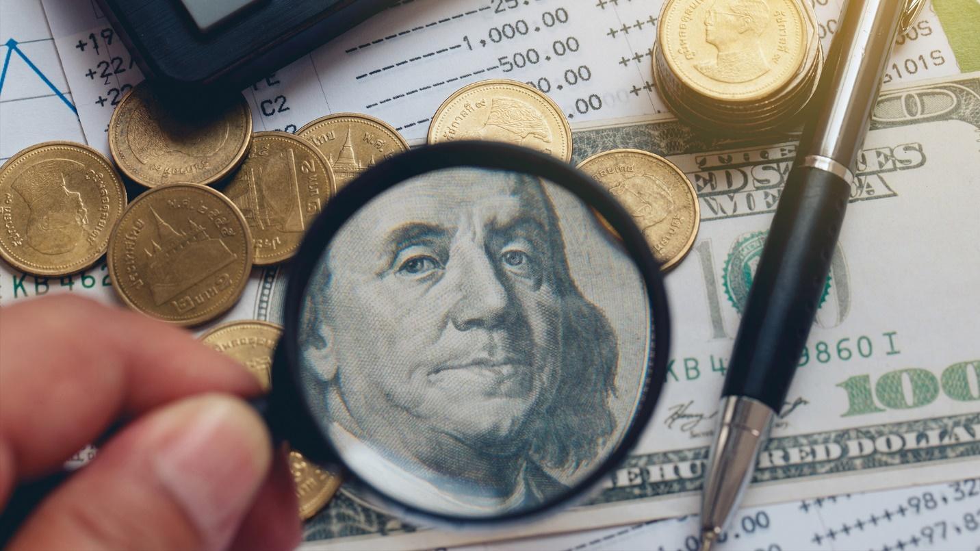 A person holds a magnifying glass up to a hundred-dollar bill and a pile of coins in a section about the Act 60 tax incentive program in Puerto Rico. 