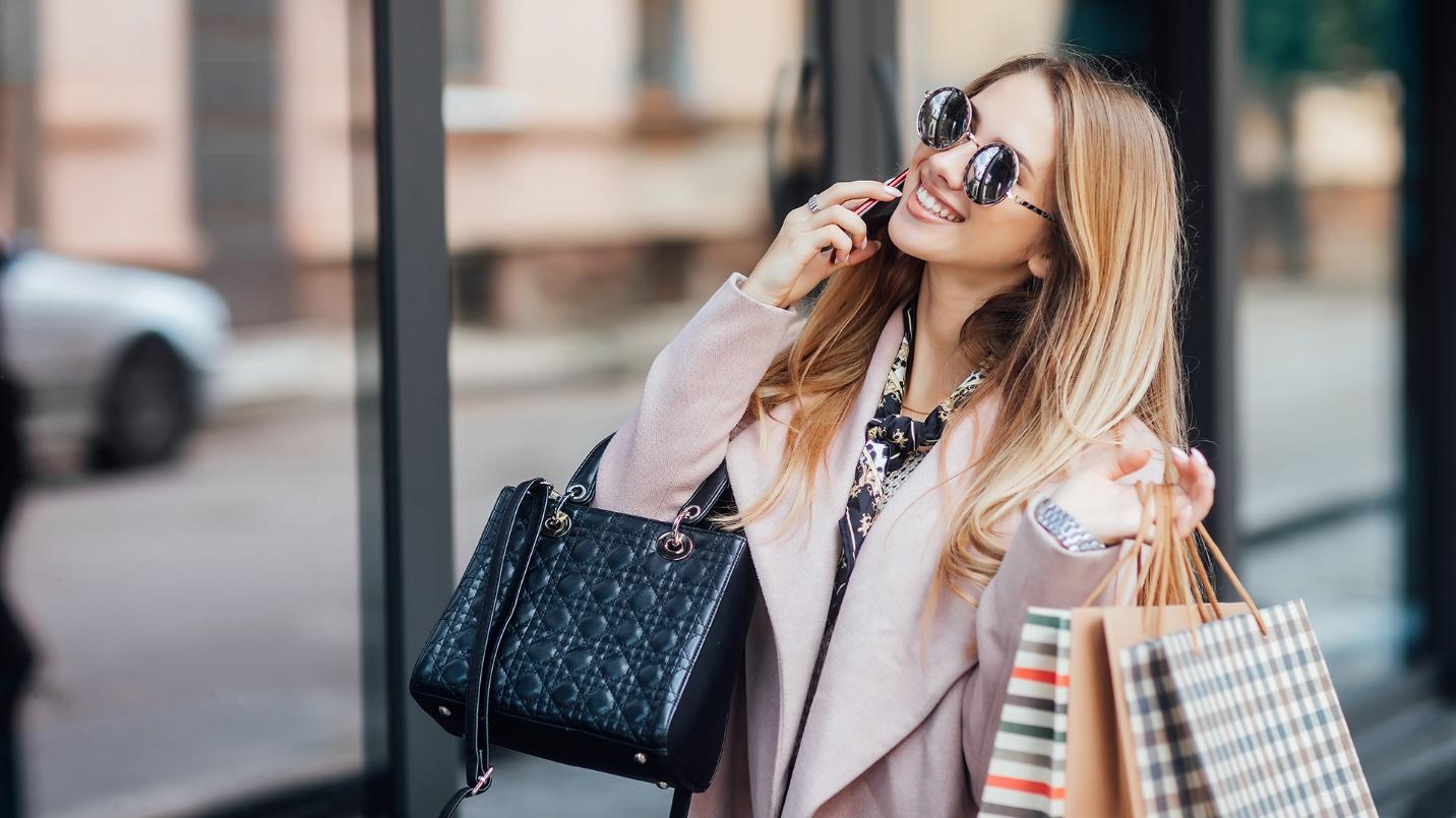 A smiling blonde woman in a pink coat and sunglasses carries bags while walking in a section about shopping in Miramar.