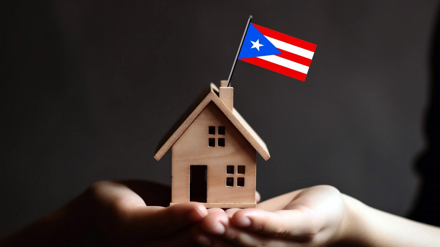 Hands hold a model wooden house topped with a Puerto Rican flag in a section previewing real estate in Miramar, Puerto Rico.