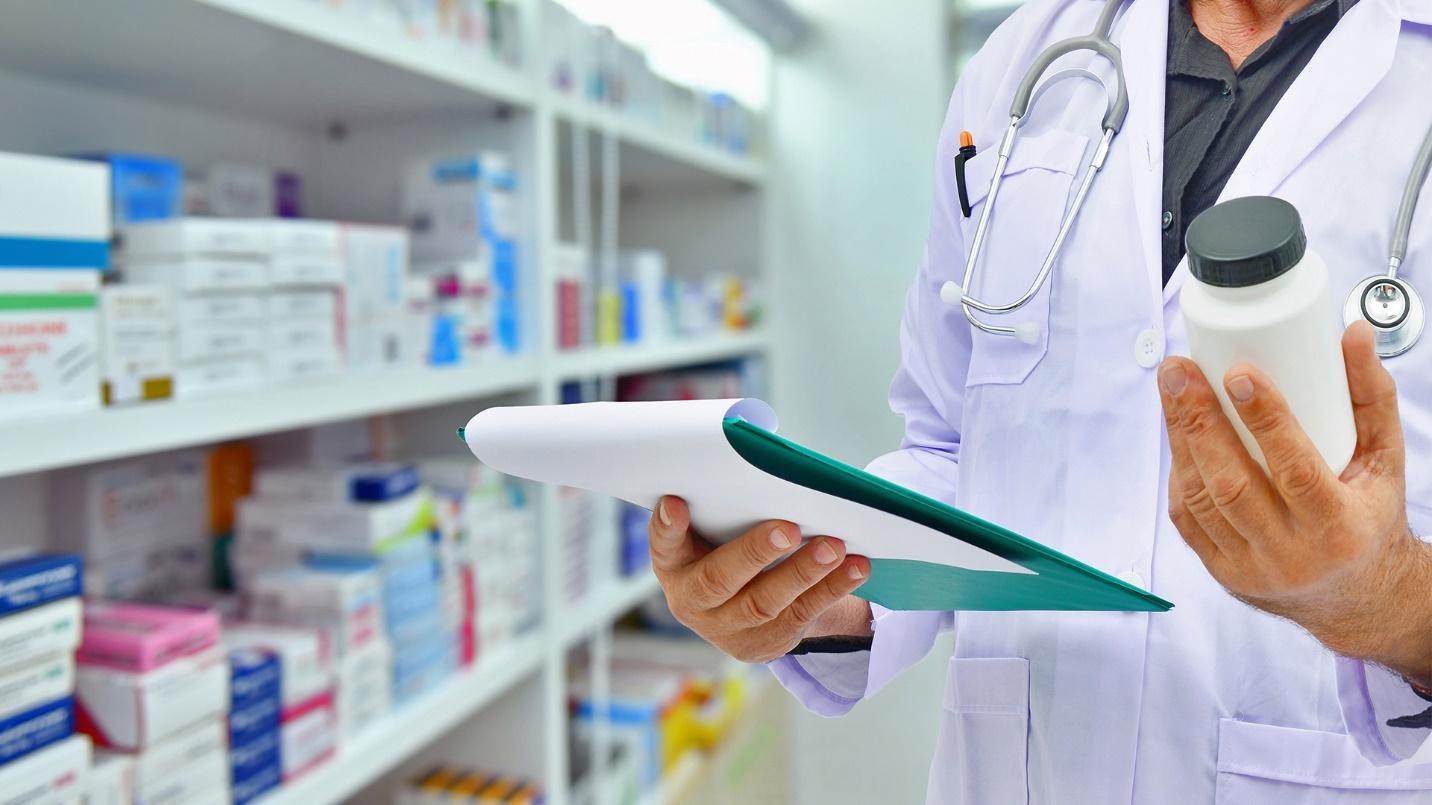 A doctor wearing a white coat holds a clipboard and bottle by shelves of medicine in a section about health insurance.
