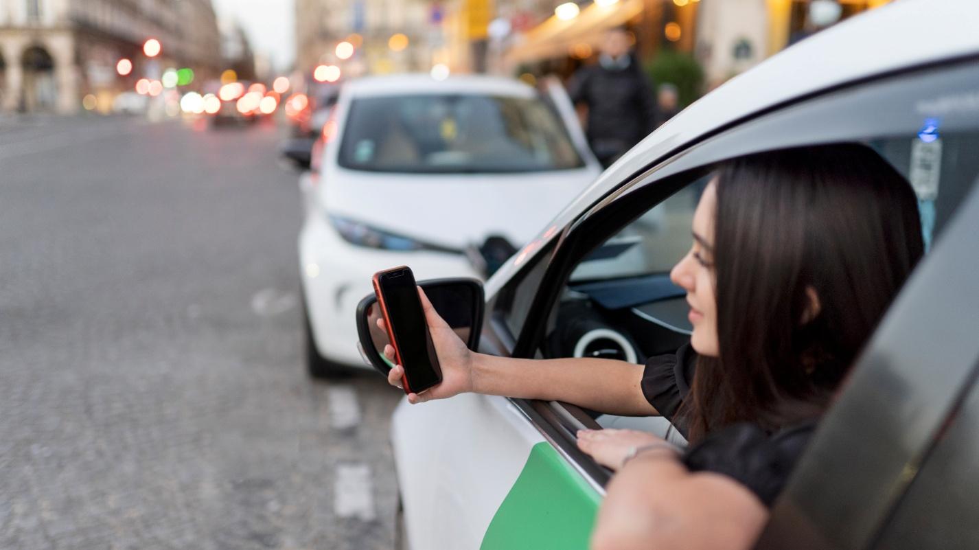 Woman using a ride-hailing app on her smartphone while leaning out of a parked car in a busy urban street, representing Uber use in Puerto Rico.