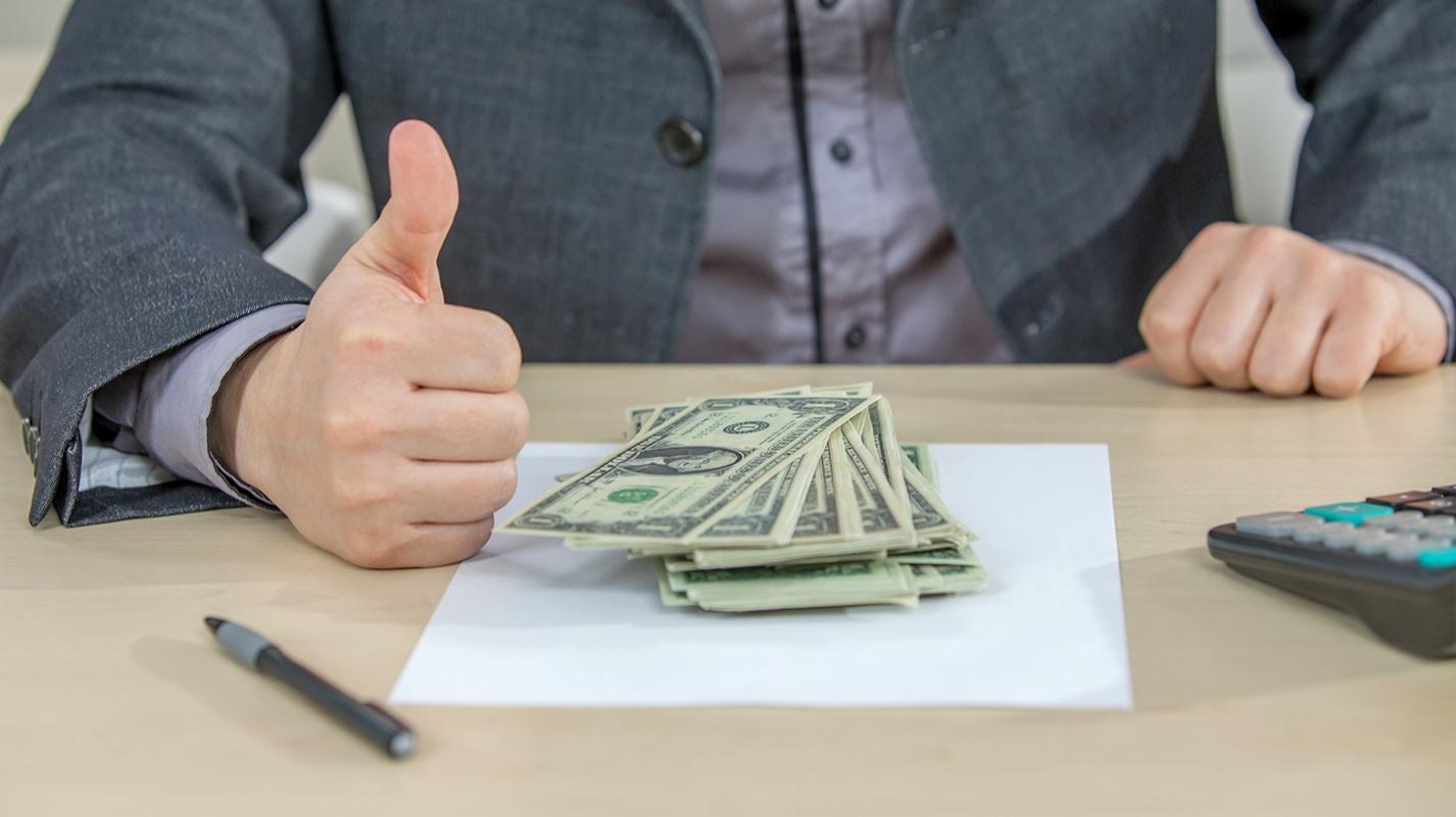 Business professional giving a thumbs up with a stack of U.S. dollar bills on a desk, indicating positive financial outcomes from effective tax strategies with the help of the PRelocate team.